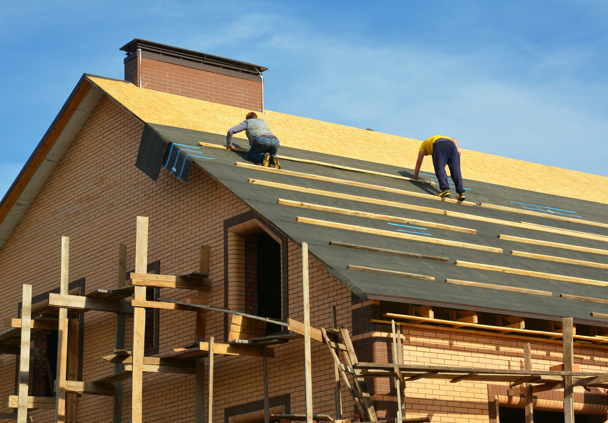 Roofing contractors are installing underlayment, waterproofing membrane on a roof deck, plywood sheathing of a large brick house under construction with scaffolding. Experienced roofing contractors evaluating roof condition for hurricane readiness – All Phase Roofing